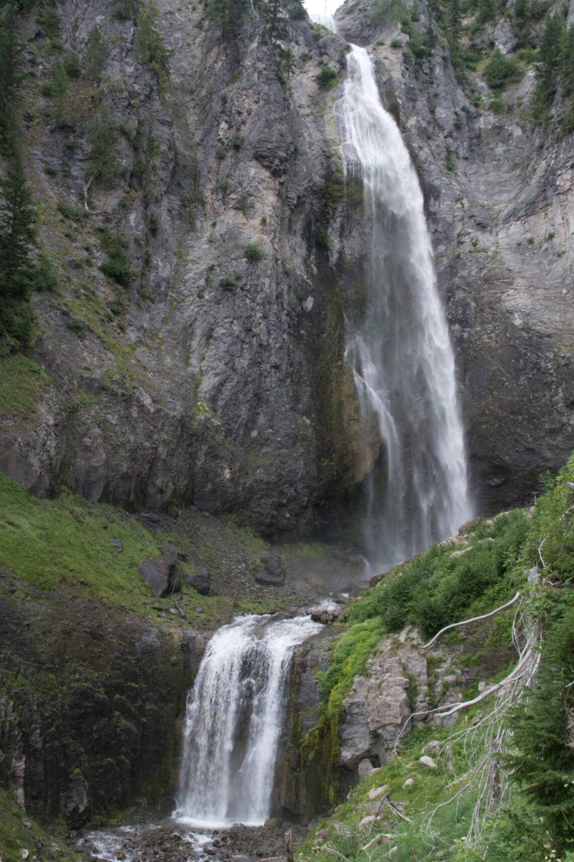 Two-tier waterfall flowing off a cliff and also over a lower drop coated in green foliage.